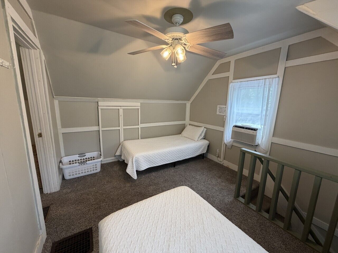 Un dormitorio con una cama individual, ventilador de techo, ventana con calentador y cesto de ropa.