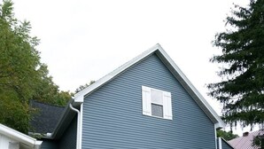 Uma casa de dois andares com varanda da frente, um pequeno jardim frontal e uma porta de garagem visível.