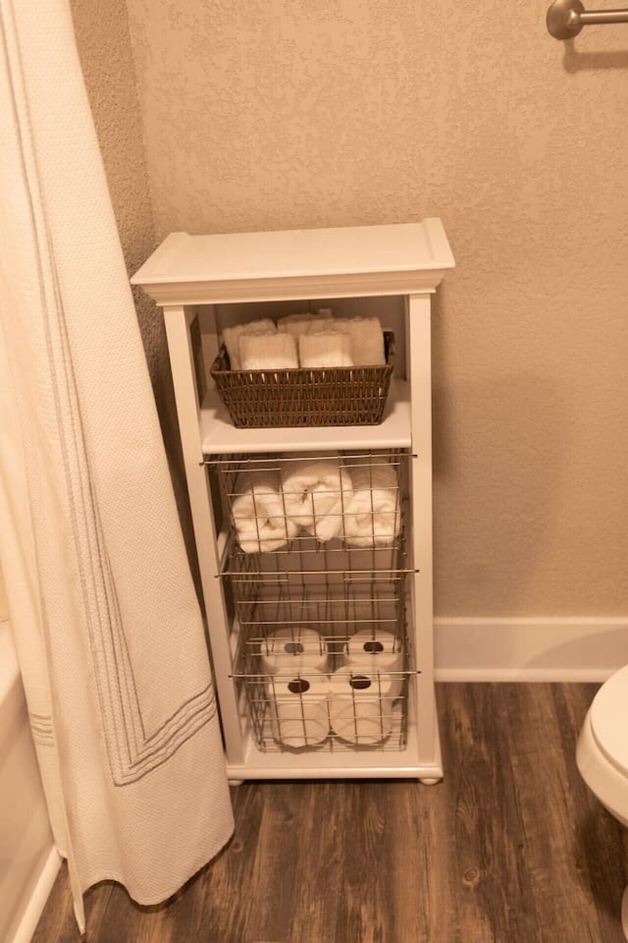A bathroom with a white cabinet containing a wicker basket, a wire shelf with towels, and a shower curtain.