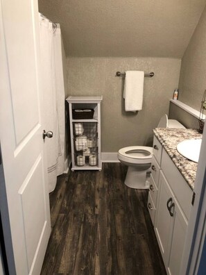 A compact bathroom with a toilet, a small shelf with baskets, and a towel hanging on the wall. - Spacious Rustic Downtown Market St 1 Bedroom Apt, Steps to Honeywell & Dining (Wabash)