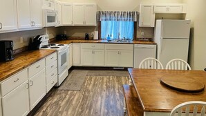 A compact kitchen with white cabinets, a microwave, stove, and refrigerator. There is a wooden dining table with chairs and a window with curtains.