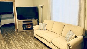 A living room with a beige sofa, a flat-screen TV, a fireplace, and a large window with sheer curtains.