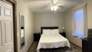 A bedroom with a bed, a ceiling fan, a nightstand, a dresser, and a window with curtains.