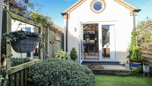A small, single-story house with a white exterior and a brown roof. It features a circular window above the door and a small porch with two chairs. - The Garden Cottage (Hinckley)