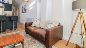 A spacious living room with a brown leather sofa, a wooden coffee table, and a floor lamp. There is a kitchen area with dark cabinets and a window above the counter. - The Garden Cottage (Hinckley)