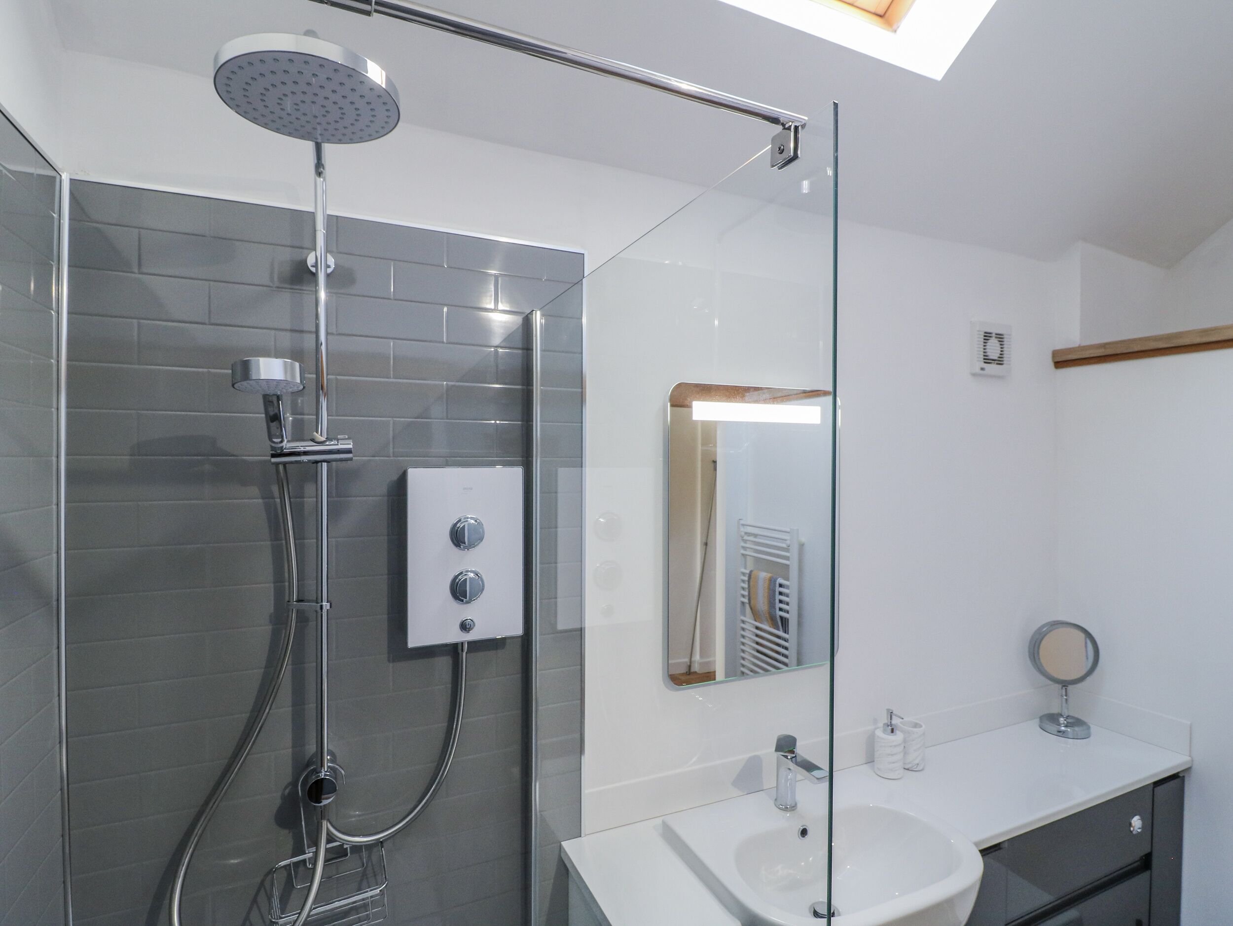 A modern bathroom with a glass shower enclosure, a wall-mounted control panel, a vanity with a sink, and a mirror.