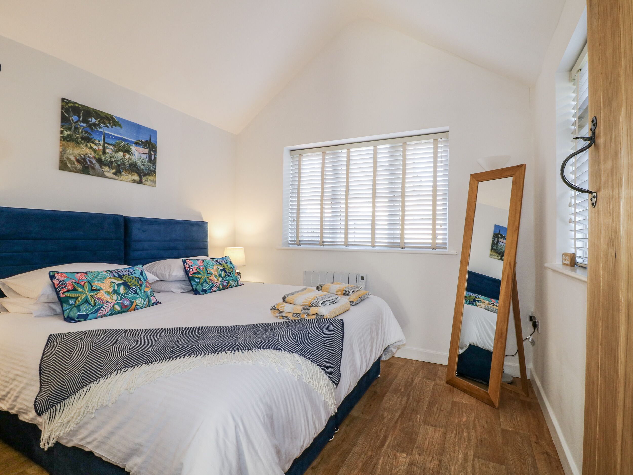 A bedroom with a large bed, a mirror, a window with blinds, and a painting on the wall.