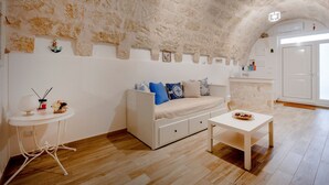 Een kamer met een stenen muur, een witte bank met blauwe kussens, een klein rond tafeltje met een schaal, en een witte kast.