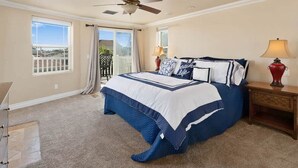 Un dormitorio con cama, ventilador de techo, mesita de noche con lámpara y vistas al exterior.