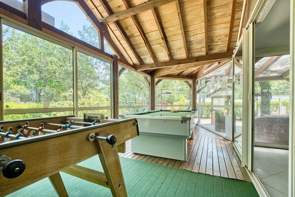 A pool table, foosball table, and large windows in a covered area.