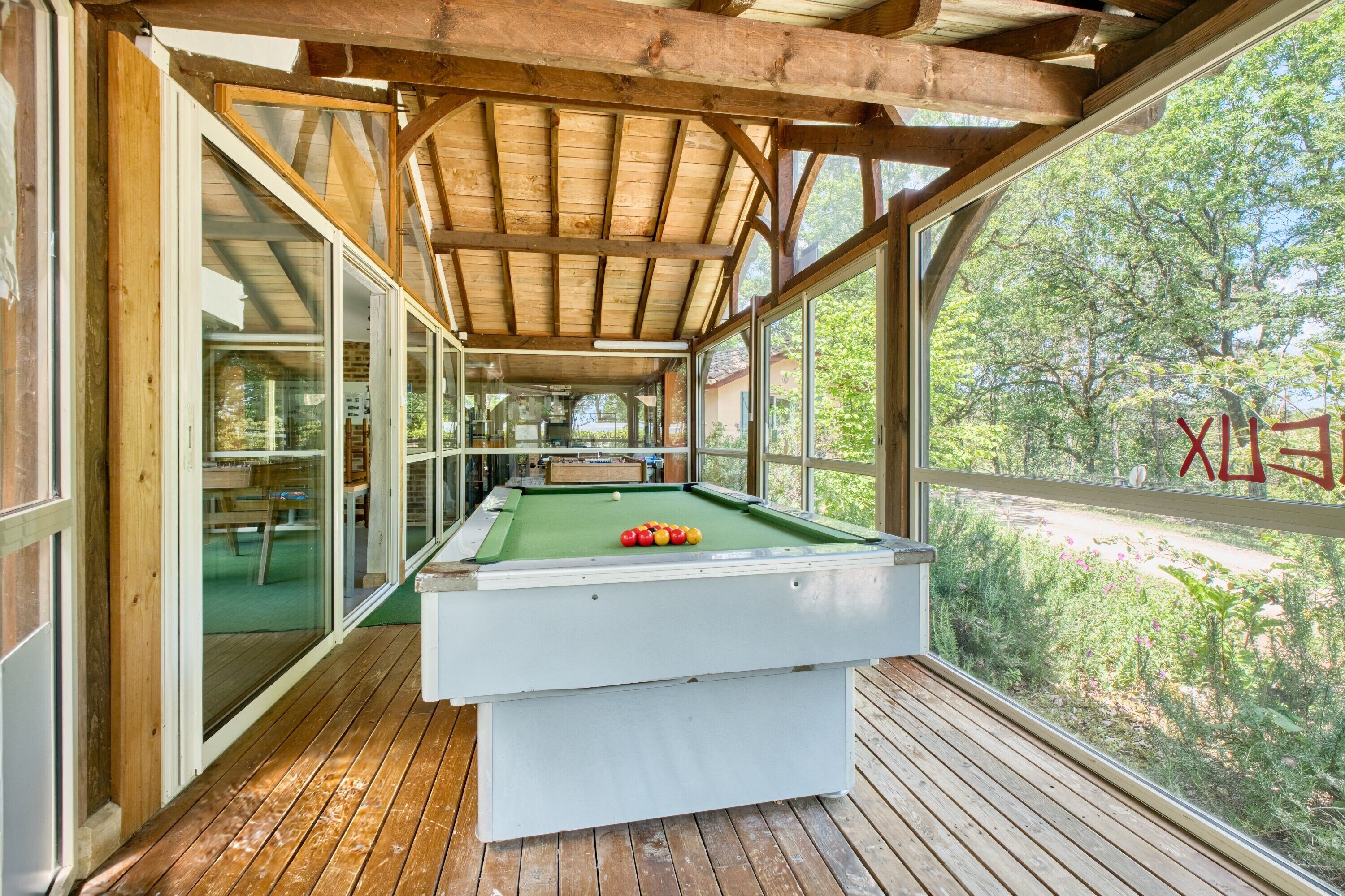 A pool table in a covered area with wooden beams and glass walls.