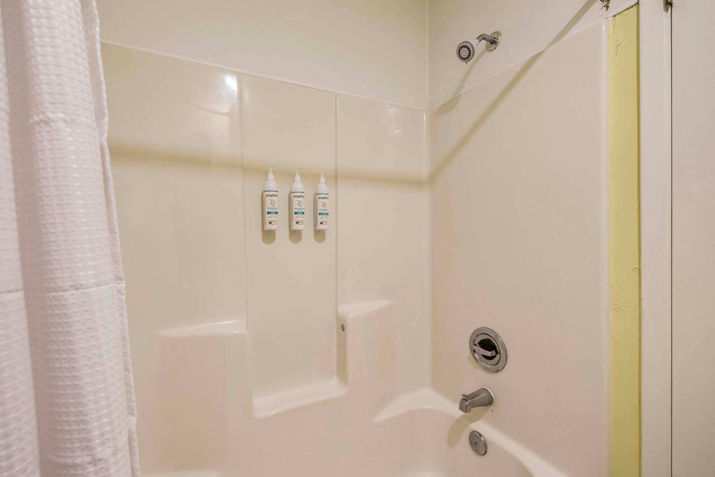 A bathroom with a white tiled wall, a shower area, and three bottles of shampoo on the wall.