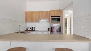 A modern kitchen with wooden cabinets, a central island with stools, a built-in oven, and a microwave. - Direct Beachfront Townhouse (Malua Bay)