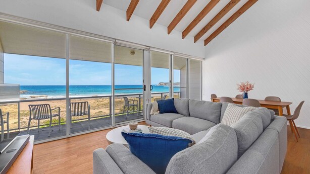 A modern living room with a large sliding glass door leading to a balcony overlooking a beach. - Direct Beachfront Townhouse (Malua Bay)