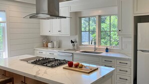 A modern kitchen with white cabinets, a large island with a gas stove, a sink, and a refrigerator. There are wooden stools at the island and a window providing natural light. - Away - Ocracoke Island (Ocracoke)
