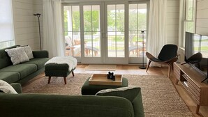 A living room with a green sofa, a wooden coffee table, and a TV stand. There is a sliding glass door leading to an outdoor patio area. - Away - Ocracoke Island (Ocracoke)
