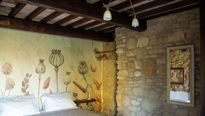 Une chambre avec un mur en pierre, un lit avec une literie à rayures, une lampe suspendue et un miroir fixé au mur.