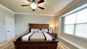 A bedroom with a wooden bed, a ceiling fan, a nightstand, a lamp, and a window with blinds. - Cozy Family Retreat (Rosenberg)