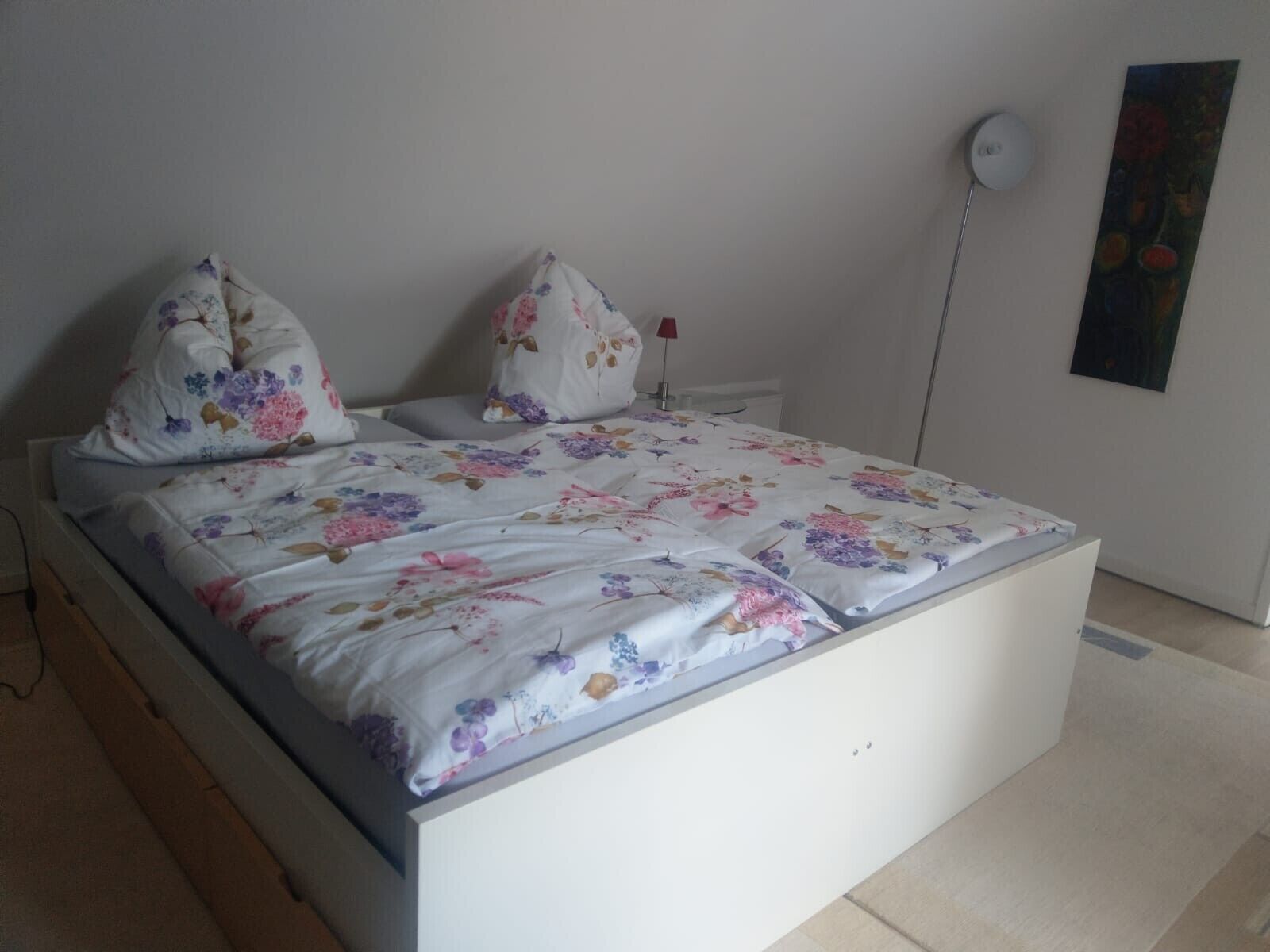 Ein Bett mit floralem Bettzeug, ein Nachttisch, eine Lampe und ein Gemälde an der Wand.