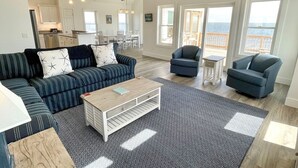 A spacious living room with a sofa, armchairs, and a coffee table. There is a kitchen in the background with white cabinets and a dining area. Large windows offer a view of the ocean. - Sundance - Ocracoke Island (Ocracoke)