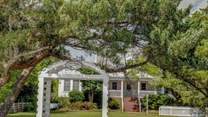 Um gazebo branco com uma placa que diz 'Wehbe House 1887' em frente a uma casa com varanda e cerca branca de piquete.