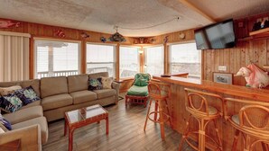 Un interior de cabaña acogedor con un sofá, una zona de comedor con sillas de mimbre, una barra y un televisor montado en la pared.