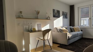 Ein modernes Wohnzimmer mit grauem Sofa, einem Holztisch und einem Schreibtisch mit Stuhl. Zwei Wandregale zeigen Dekorationsobjekte, und ein Fenster ist mit Vorhängen drapiert.