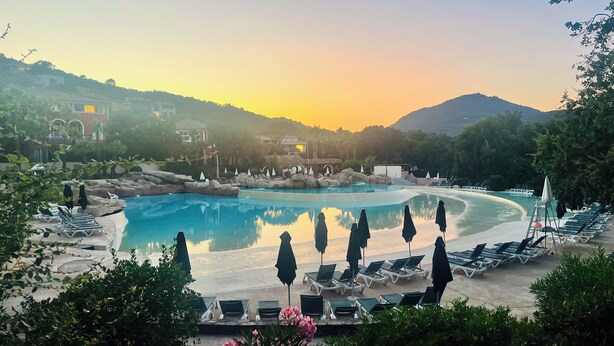 Piscina de resort com espreguiçadeiras e guarda-sóis, cercada por vegetação e colinas.