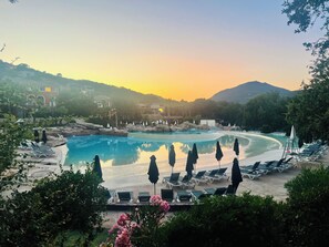 Une piscine de complexe hôtelier, dotée de chaises longues et de parasols, entourée de verdure et de collines.