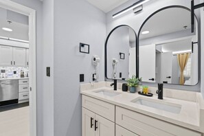 Une salle de bain moderne avec un double vasque, deux sèche-cheveux, un grand miroir et une plante en pot.