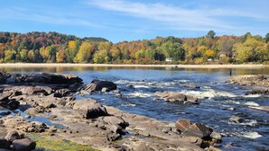 A rocky shoreline with a river, a forested hill in the background, and a house near the water's edge. - Little House, Rivière Rouge waterfront, 7 minutes from Mont-Tremblant town. (Brébeuf)