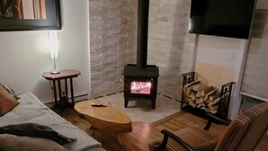 A cozy living room with a wooden floor, a sofa, a chair, a small table, a TV mounted on the wall, and a wood-burning stove. - Little House, Rivière Rouge waterfront, 7 minutes from Mont-Tremblant town. (Brébeuf)