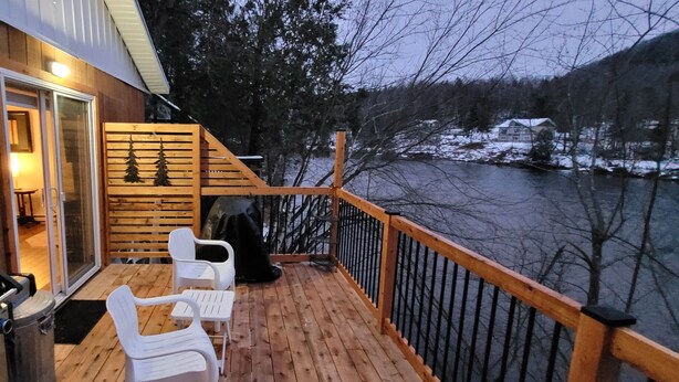 A wooden deck with a grill, white chairs, and a view of a river and forest. - Little House, Rivière Rouge waterfront, 7 minutes from Mont-Tremblant town. (Brébeuf)