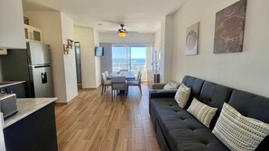 Un apartament modern amb cuina, menjador i sala d'estar. Té un finestral gran amb vistes, un ventilador de sostre i una televisió de pantalla plana.