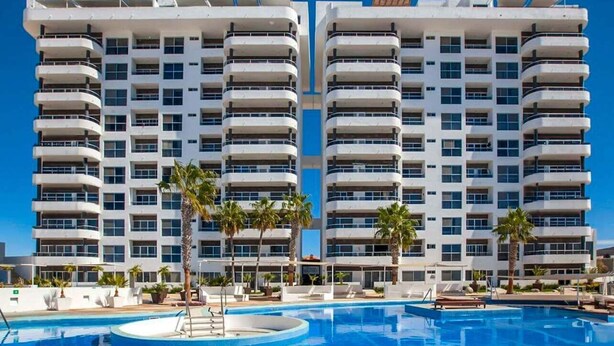 Un hotel modern amb una piscina central, múltiples balcons i palmeres.