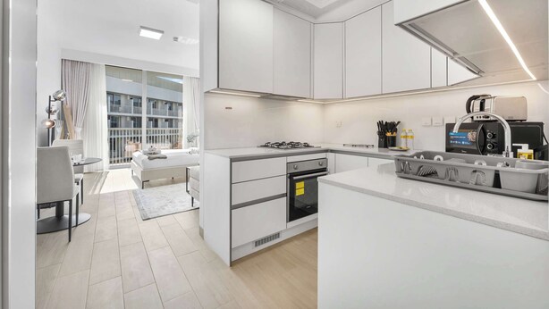 A modern kitchen with white cabinetry, a built-in oven, and a large island with a sink. - Stayshort - Contemporary Studio With Open Layout in JVC (Dubai)