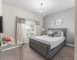 A bedroom with a grey upholstered bed, a chandelier, a bedside table with a lamp, and a window with curtains. - 5BR Home w/ Private Pool at Storey Lake (kissimmee)