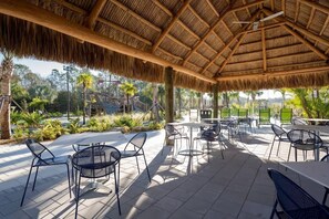 An outdoor patio area with round tables and chairs, a thatched roof, and a view of palm trees and greenery. - 5BR Home w/ Private Pool at Storey Lake (kissimmee)