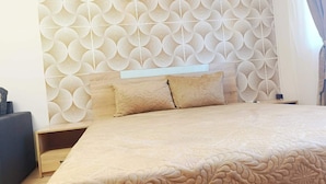 Un dormitorio con papel tapiz estampado, una cama con edredón y almohadas color beige, una mesita de noche y una mesilla.