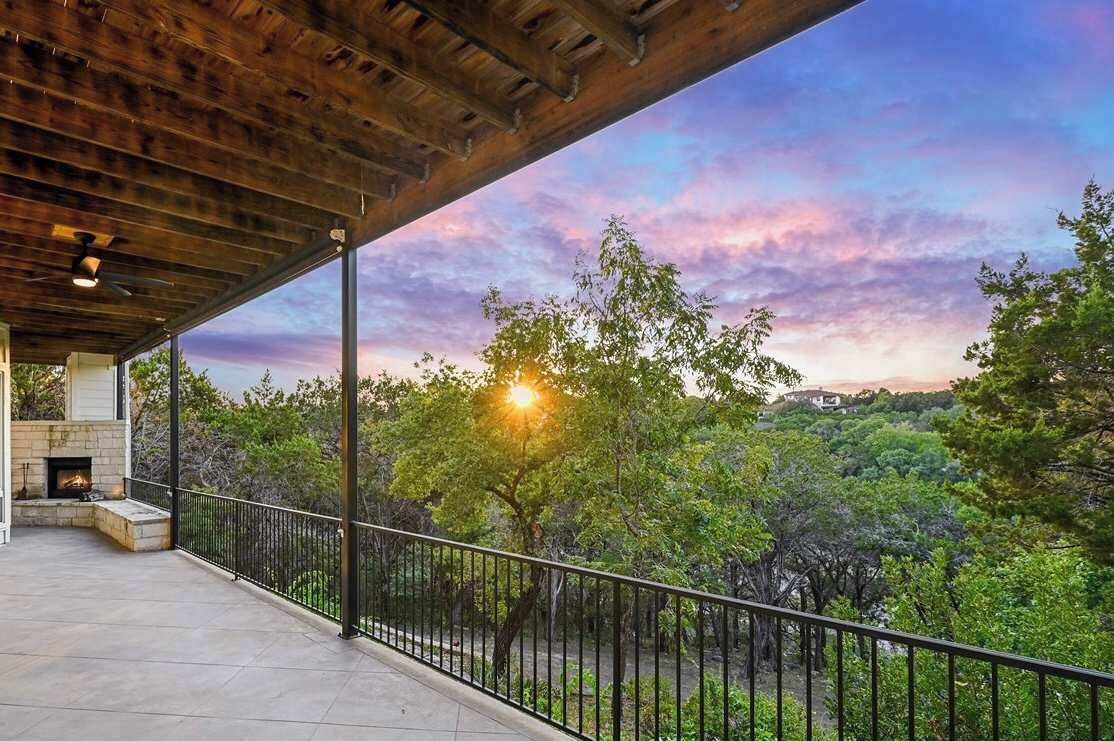 A covered outdoor area with a fireplace, a metal railing, and a view of trees and a sunset.
