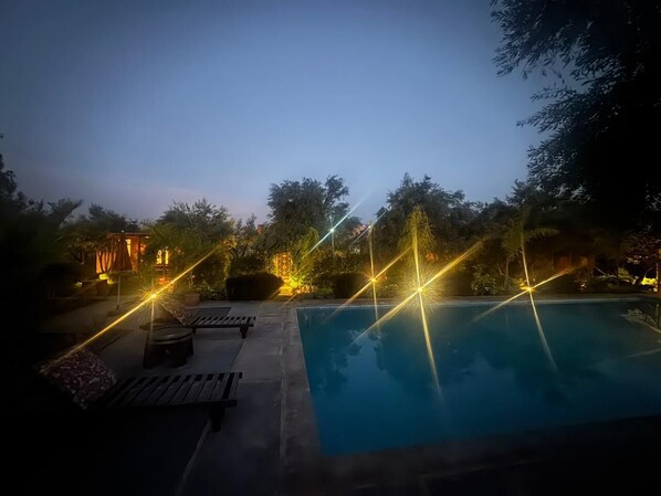 Zona de piscina nocturna con vista despejada, rodeada de árboles y un edificio con iluminación cálida.