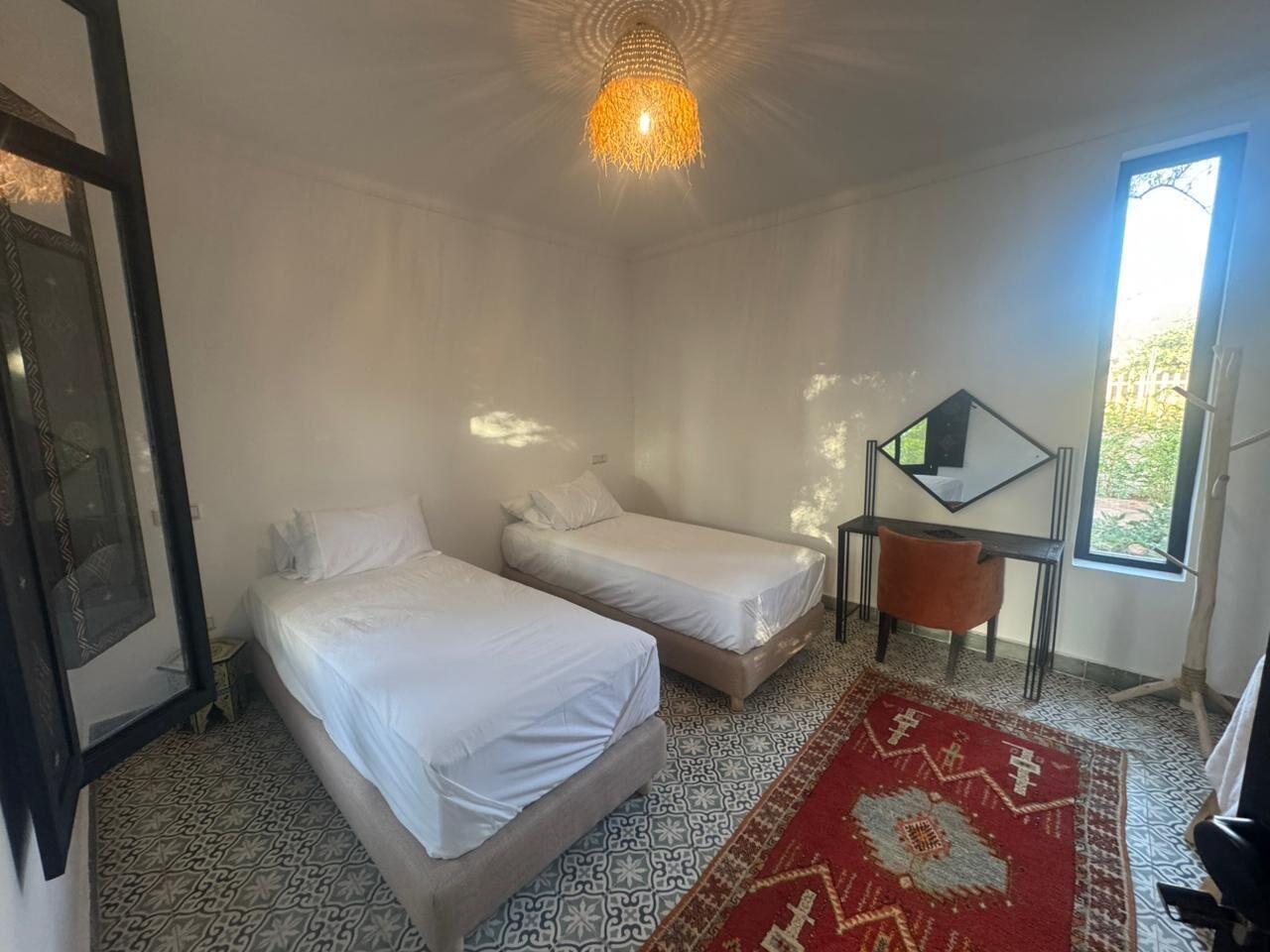 Ett rum med två sängar, en röd, mönstrad matta, en hängande lampa och ett fönster med utsikt över grönområden.