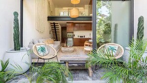 Een moderne woonkamer met een bank, twee rieten stoelen, een salontafel, en een keuken zichtbaar door een glazen deur.