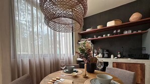 A dining area with a round wooden table, a woven placemat, and a vase with dried plants. There are wooden chairs, a hanging light fixture, and a kitchen area with shelves in the background. - Exquisitely Decorated 2 BD Retreat in Tulum (Tulum)