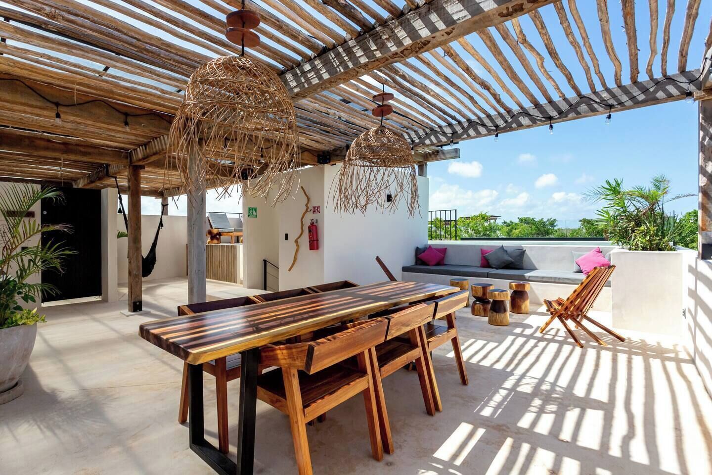 An outdoor patio with a wooden table and chairs, a hanging light fixture, and a cushioned seating area with a view of greenery.