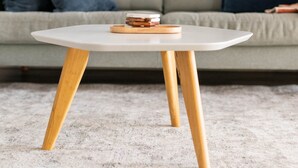 Ein runder Tisch mit Holzbeinen und einer weißen Tischplatte, darauf einige Bücher, vor einer grauen Couch mit blauen Kissen.