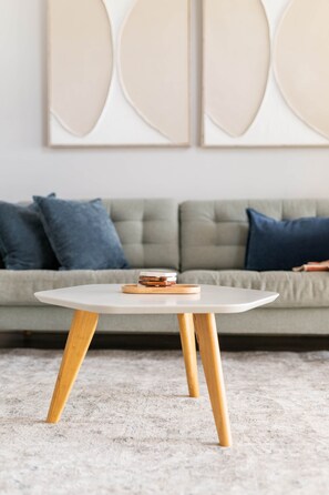 Ein runder Tisch mit Holzbeinen und einer weißen Tischplatte, darauf einige Bücher, vor einer grauen Couch mit blauen Kissen.