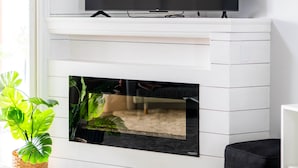 Ein modernes Wohnzimmer mit einem Wand-Fernseher, der 'NETFLIX' in roten Buchstaben anzeigt, einem Kamin darunter und einer Blumentopf auf der linken Seite.