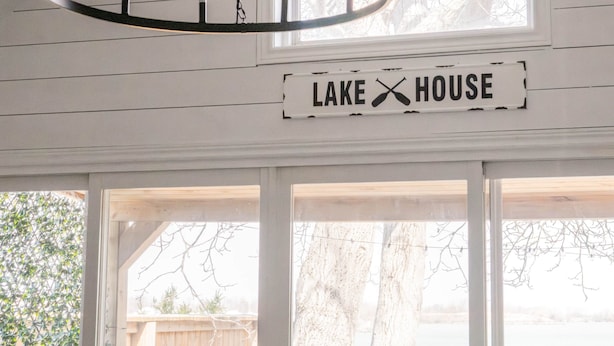 Ein Chandelier, ein Schild mit der Aufschrift 'LAKE HOUSE' und Blick auf einen Garten mit Holzzaun und Bäumen.