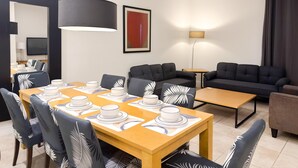 Una sala de estar moderna con comedor, que incluye una mesa de madera para cuatro personas, sofás oscuros y una lámpara colgante negra de gran tamaño.